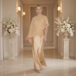 This elegant ivory silk dress features a cowl neckline and detachable sheer cape, creating a refined, graceful look ideal for bridal parties, weddings, and formal occasions. Its flowing fabric and asymmetrical cape hem offer a stylish balance of elegance and comfort. Perfect as a bridesmaid gift or wedding guest outfit, this dress radiates timeless sophistication and is a wonderful choice for any special event or celebration. Bridal gifting made luxurious.