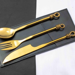 May include: A gold-colored flatware set including a spoon, fork, and knife, resting on a black leather-like placemat. The handles have a looped design. The background is split between black and white.