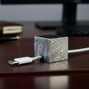 Puede incluir: Un organizador de cables cuadrado de metal estampado con un cable blanco insertado. El organizador tiene un diseño único y arremolinado en tonos grises y negros. El cable está conectado a un conector USB-C blanco.