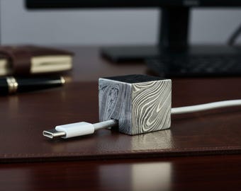 May include: A square, patterned metal cable organizer with a white cable inserted. The organizer has a unique, swirling design in shades of gray and black. The cable is connected to a white USB-C connector.