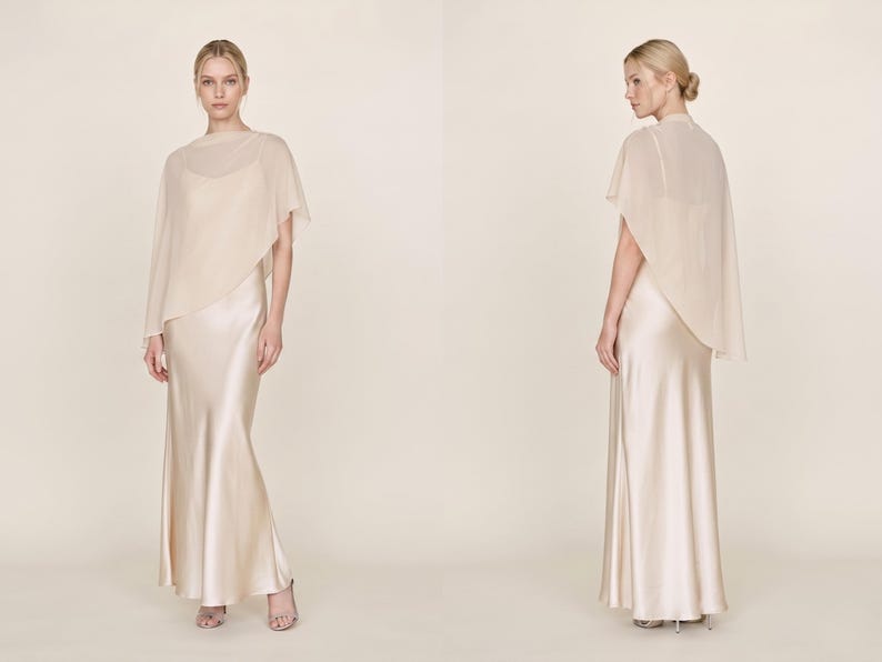 This ivory silk gown with a detachable sheer cape is the epitome of bridal elegance. Featuring a flattering cowl neckline and asymmetrical cape hem, it makes a stunning addition to any formal occasion, from weddings to bridal showers. Ideal as a bridal gift or a bridesmaid dress, this gown embodies sophistication and timeless charm, making it the perfect choice for those looking for an exquisite bridal party outfit or a thoughtful fashion gift.