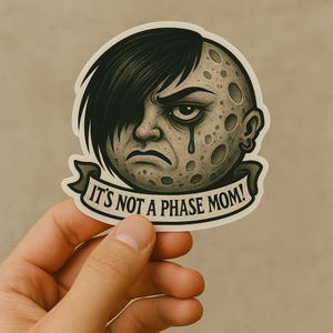 以下が含まれることがあります： 黒い髪と涙を持つ月の顔のキャラクターが描かれたステッカーで、「IT'S NOT A PHASE MOM！」というテキストがバナーに表示されています。キャラクターは悲しい表情をしています。
