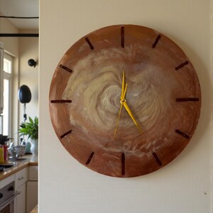 May include: A round wall clock with a brown wooden frame and a swirling cream and brown center. The clock has yellow hands and dark brown markers for the hours. The clock is mounted on a light beige wall.