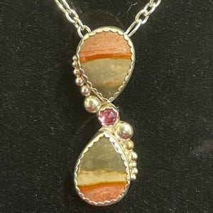 Puede incluir: Un collar de plata con un colgante con dos piedras en forma de lágrima en tonos rojos, naranjas y grises. Una pequeña gema rosa y cuentas de plata acentúan el colgante. La cadena del collar es una delicada cadena de plata.