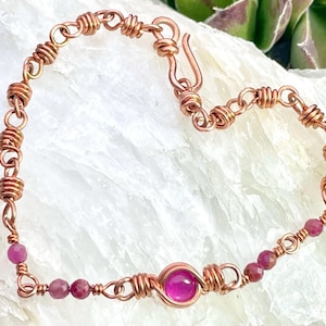 May include: A handcrafted copper bracelet featuring a series of wrapped wire links and small, round, pink gemstones. The bracelet includes a larger, central pink gemstone and a hook clasp. The jewelry is displayed on a white surface.