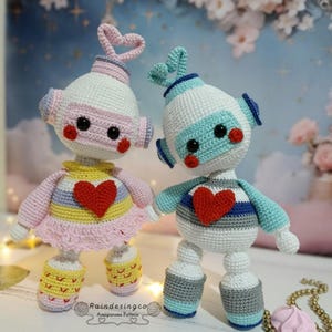 Puede incluir: Dos muñecas de ganchillo hechas a mano. Una muñeca es rosa y blanca con un adorno para la cabeza en forma de corazón y un corazón rojo en el pecho. La otra muñeca es azul y blanca con un adorno para la cabeza en forma de corazón y un corazón rojo en el pecho. Ambas muñecas tienen ojos negros y mejillas rojas.