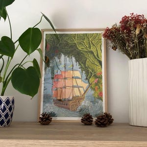 Op de afbeelding: Een ingelijste prent van een zeilschip dat door een weelderig, groen en bloemrijk landschap vaart. De zeilen van het schip zijn een mix van oranje en roze. Het kunstwerk wordt tentoongesteld op een houten plank met decoratieve dennenappels en een potplant.