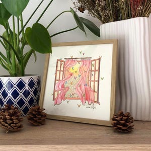 Peut inclure: Une impression encadrée à l'aquarelle représente une fenêtre ouverte avec des rideaux roses. Des papillons volent autour de la fenêtre, avec un ciel jaune et orange vif visible. L'œuvre est dans un cadre en bois clair, avec une plante et des pommes de pin au premier plan.