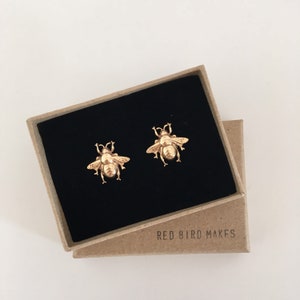 Op de afbeelding: Een paar gouden bijen oorbellen in een klein bruin doosje met de tekst "RED BIRD MAKES" op de bodem.