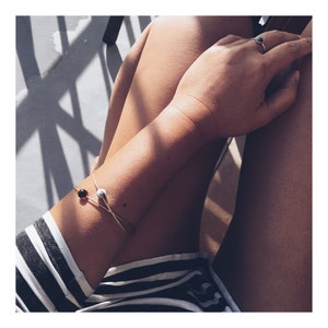 May include: A gold bracelet with two small round beads, one brown and one white, on a person's wrist. The bracelet is on the left wrist and the person is wearing a black and white striped shirt.
