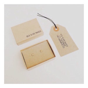 May include: A small, brown box with the text "RED BIRD MAKES" on the top. The box contains two gold, butterfly-shaped earrings. A brown tag with the text "RED BIRD MAKES FACEBOOK INSTAGRAM ETSY" is attached to the box with a black string.