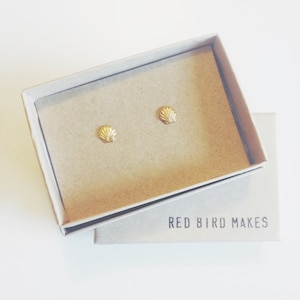 Könnte beinhalten: Ein Paar goldene Muschel-Ohrringe in einer kleinen Pappbox mit dem Text "RED BIRD MAKES" auf dem Boden der Box.