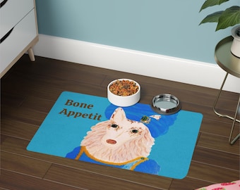 May include: A blue pet food mat with the words "Bone Appetit" and a whimsical illustration of a dog wearing a blue hat and jacket. A white ceramic bowl and a silver bowl are on the mat.