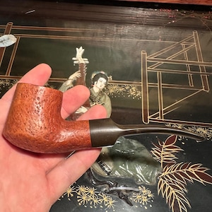 Peut inclure: Une pipe à tabac marron avec un foyer texturé et une tige sombre. La pipe est tenue dans une main, avec une surface noire décorative en arrière-plan.