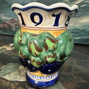 May include: A ceramic vase with a scalloped rim, featuring the year "1918" in navy blue. The vase has a green, brown, and blue design, with a bulbous midsection and a decorative base. The background is blurred.