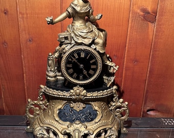 Könnte beinhalten: Eine antike, goldfarbene Uhr mit einer sitzenden Frauenfigur auf einem schwarzen Zifferblatt mit römischen Ziffern. Die Uhr hat eine aufwendige Detaillierung und steht auf einem dekorativen Sockel. Der Hintergrund ist eine Holzwand.