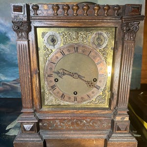 May include: Vintage wooden mantel clock with a brass face. The clock has Roman numerals, decorative hands, and two smaller dials. The wooden frame features intricate carvings and a dark brown finish. A classic antique piece.