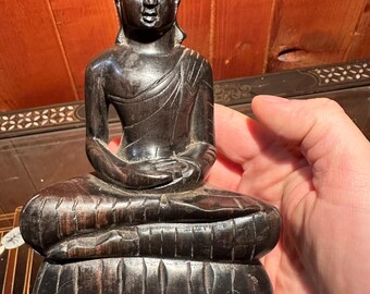Op de afbeelding: Een donkerbruin houten Boeddha-beeld in een zittende meditatiehouding. Het beeld heeft gedetailleerde gravures en wordt door een hand vastgehouden. De Boeddha zit op een lotusbloem basis, met de handen in de schoot.