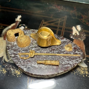 May include: A selection of gold-toned miniature military regalia, including a bicorne hat, epaulettes, a sword, a telescope, and a sunburst emblem, displayed on a marble-effect surface. These items are likely decorative or collectible.