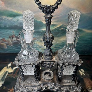 May include: An antique silver-toned cruet set with two glass bottles, each with a diamond-cut stopper. The bottles sit in ornate holders, connected by a handle. The set is on a decorative base.