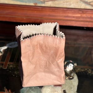 May include: A small, ceramic paper bag-shaped container in a muted pink hue. The top edge features a decorative, zig-zag design. The interior is a light beige color. The container sits on a dark, patterned surface.