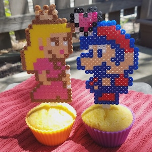 Puede incluir: Dos cupcakes decorados con figuras de pixel art de una princesa y un personaje de videojuego. La princesa lleva un vestido rosa y una corona dorada. El personaje de videojuego lleva un sombrero rojo y un mono azul.