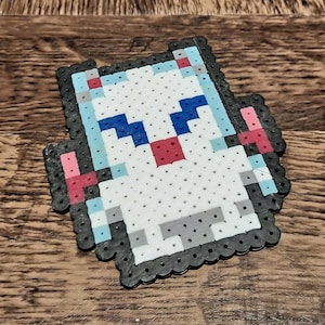May include: A pixelated cat design made from colorful plastic beads. The cat features a white body, blue eyes, and a red nose. The design is outlined in black beads and sits on a wooden surface.