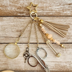 May include: A gold-toned keychain with a star-shaped charm, holding various charms including a magnifying glass, small scissors, a key, a tassel, and a beaded strand. A small wooden bowl with spools of thread is in the lower left corner.