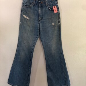 May include: Faded blue denim bell-bottom jeans with distressed details, including a rip on the left leg. The jeans hang from a wooden hanger. A pink tag is attached to the front pocket.