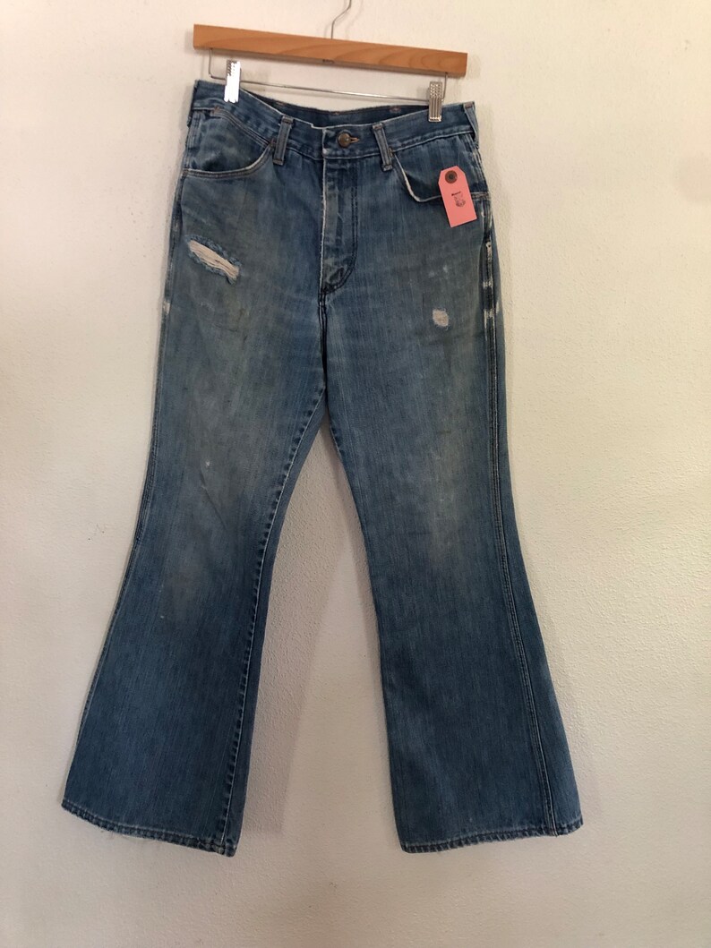 May include: Faded blue denim bell-bottom jeans with distressed details, including a rip on the left leg. The jeans hang from a wooden hanger. A pink tag is attached to the front pocket.