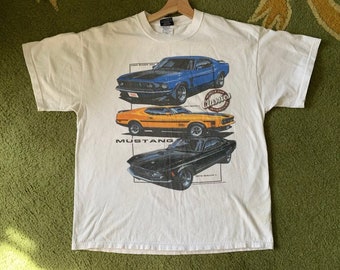 Op de afbeelding: Wit t-shirt met illustraties van drie klassieke Mustang-auto's in blauw, oranje en zwart. Het shirt heeft de tekst "Mustang" en "Muscle Car Classics" erop gedrukt. Het t-shirt hangt aan een houten hanger.