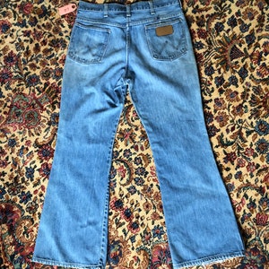 May include: Light blue denim jeans with a flared leg design. The jeans feature two back pockets, a brown leather patch with the Wrangler logo, and a classic five-pocket design. The jeans are laid flat on a patterned rug.