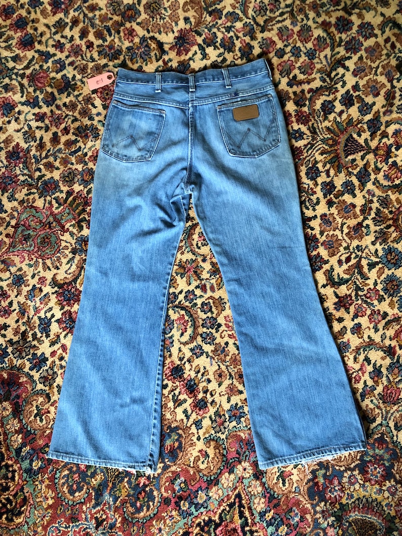 May include: Light blue denim jeans with a flared leg design. The jeans feature two back pockets, a brown leather patch with the Wrangler logo, and a classic five-pocket design. The jeans are laid flat on a patterned rug.