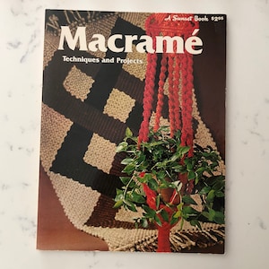 May include: A vintage book cover titled "Macramé Techniques and Projects" featuring a red macramé plant hanger holding a potted plant with green leaves. The background shows a macramé wall hanging with brown, black, and beige patterns.