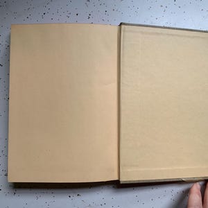 May include: An open book with tan-colored pages. The left page is fully visible, while the right page is partially visible. The book's spine is a darker color, and the pages have a slightly aged appearance. The book is on a speckled gray surface.