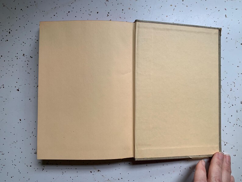 May include: An open book with tan-colored pages. The left page is fully visible, while the right page is partially visible. The book's spine is a darker color, and the pages have a slightly aged appearance. The book is on a speckled gray surface.