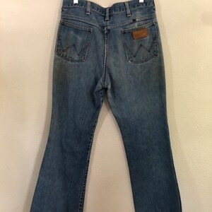 May include: Light blue denim Wrangler jeans hanging on a wooden hanger. The jeans have two back pockets, a brown leather patch, and a flared leg design. The jeans are faded and show signs of wear.