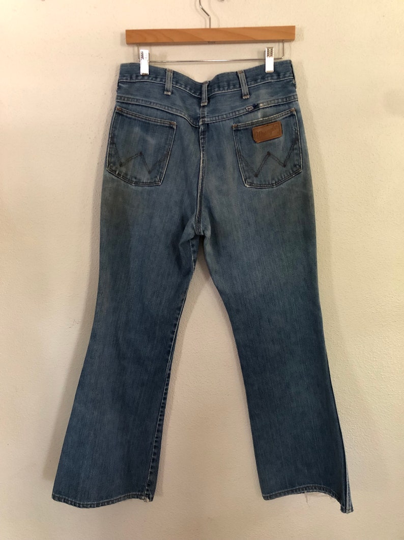 May include: Light blue denim Wrangler jeans hanging on a wooden hanger. The jeans have two back pockets, a brown leather patch, and a flared leg design. The jeans are faded and show signs of wear.