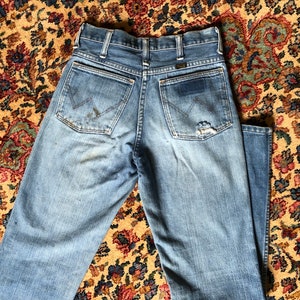 May include: Light blue denim jeans with a classic five-pocket design. The jeans show signs of wear, including a small tear on the back pocket and some discoloration. The Wrangler logo is visible on the back pocket.