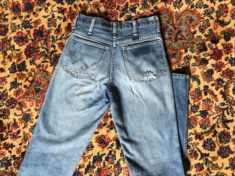 May include: Light blue denim jeans with a classic five-pocket design. The jeans show signs of wear, including a small tear on the back pocket and some discoloration. The Wrangler logo is visible on the back pocket.