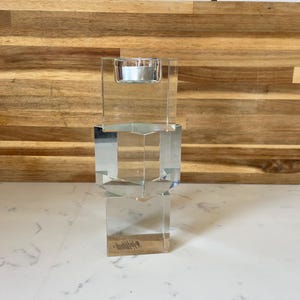 May include: A clear, geometric glass candle holder with a small lit candle. The holder has a rectangular base and a square top, with a series of geometric shapes in between. The background is a wooden surface.