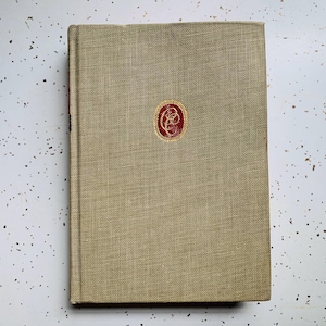 May include: A hardcover book with a tan linen cover and a red emblem in the center. The book's spine is visible on the left side. The background is a white surface with black speckles.