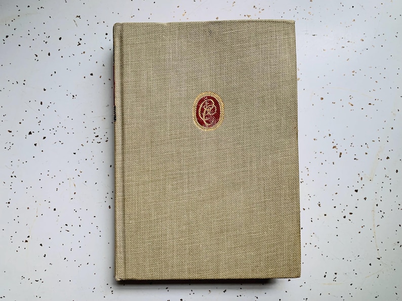 May include: A hardcover book with a tan linen cover and a red emblem in the center. The book's spine is visible on the left side. The background is a white surface with black speckles.