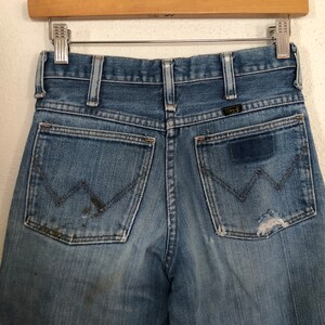May include: Close-up of vintage blue denim jeans with a classic five-pocket design. The jeans show signs of wear, including fading, a patch, and a small tear on the back pocket. The Wrangler logo is visible.