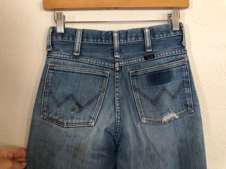 May include: Close-up of vintage blue denim jeans with a classic five-pocket design. The jeans show signs of wear, including fading, a patch, and a small tear on the back pocket. The Wrangler logo is visible.