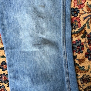May include: Close-up of light blue denim jeans with visible wear and tear. The jeans have a faded appearance with subtle distressing. The stitching is visible along the seams. The jeans are on a patterned rug.