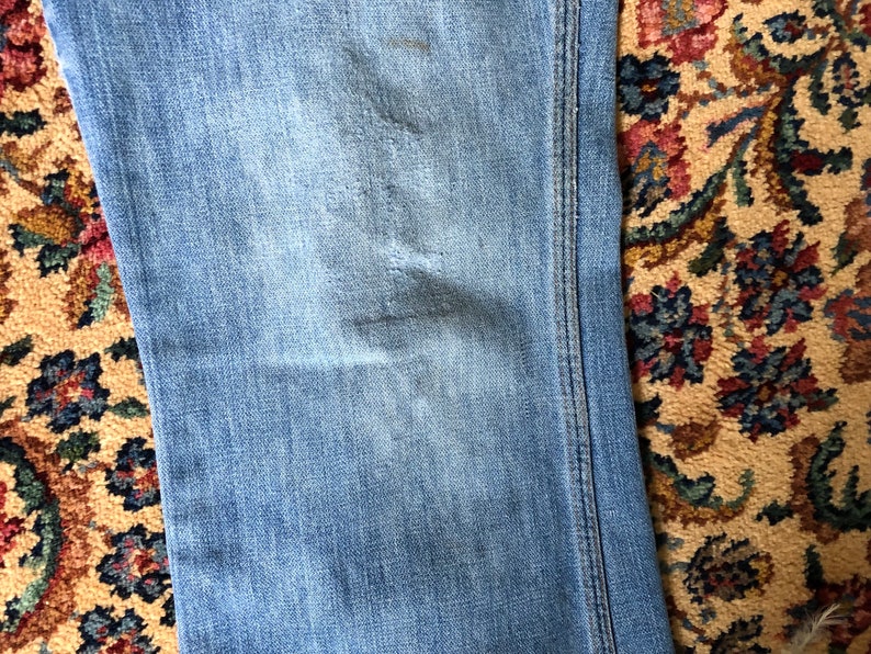 May include: Close-up of light blue denim jeans with visible wear and tear. The jeans have a faded appearance with subtle distressing. The stitching is visible along the seams. The jeans are on a patterned rug.