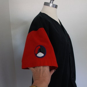 May include: Black baseball-style shirt with red sleeves. The left sleeve features a circular patch with a black and white design inside a blue circle. The shirt is button-down and displayed on a mannequin.