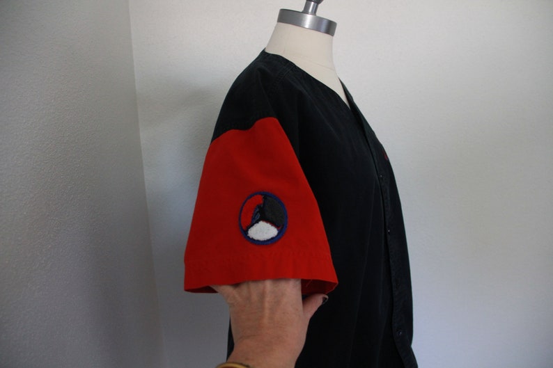 May include: Black baseball-style shirt with red sleeves. The left sleeve features a circular patch with a black and white design inside a blue circle. The shirt is button-down and displayed on a mannequin.