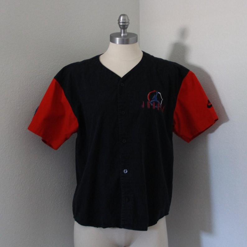 May include: Black button-down baseball jersey with red short sleeves. Features an embroidered design on the chest and a small logo on the right sleeve. The jersey is displayed on a mannequin.